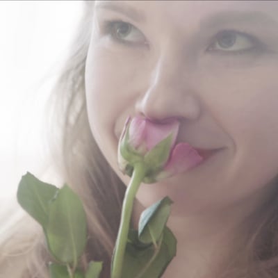 Anna-Riikka Rajanen roolihahmossaan haistelee vaaleanpunaista ruusua vaaleassa kuvassa.