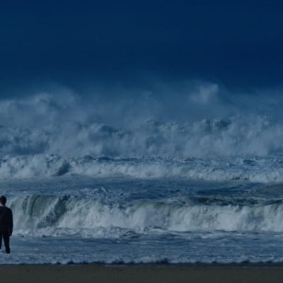 En ensam man står inför en enorm svallvåg som rullar in mot stranden.