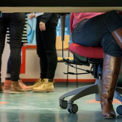 Naisen jalat ristissä työpöydän alla, taustalla  kolmen henkilön jalat. 