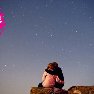 ett par som håller om varandra på en klippa och ser upp på stjärnhimmeln 