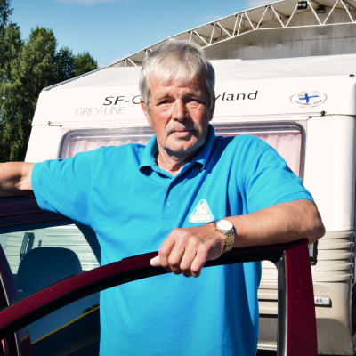 Olof Bussman i klädd en turkos pikéskjorta står lutad mot en bildörr. I bakgrunden skymtar en husvagn.