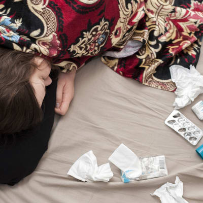 Sairastava ihminen makaa sängyllä nenäliinojen, kuumemittarin ja lääkkeiden kanssa.