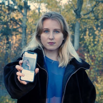 En kvinna med långt blont hår. Hon står framför en björkskog. Hon håller upp en telefon där appen Instagram är igång. Vi ser en bild av henne, där hon är lättklädd och väldigt smal, eftersom hon lider av anorexi. 