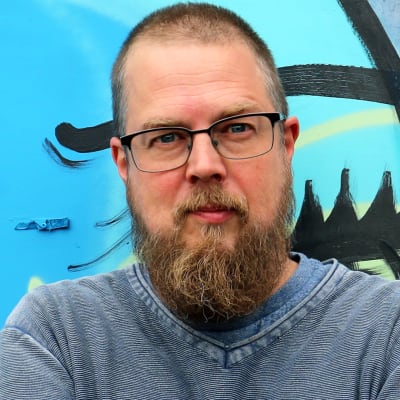 Kuvassa on kirjailija Tuomas Kyrö purkutuomion saaneen Kaurialan yläasteen, graffitin peittämän, seinän edessä.  