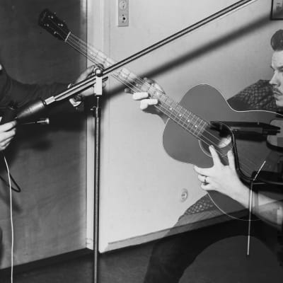 Editoidussa kuvassa toimittaja Martti Silvennoinen, joka tutustuu ääntä kaukaa ottavaan orumikrofoniin v.1957 ja häntä vastapäätä on kitaraa soittava mies osin pikselöitynä v.2019