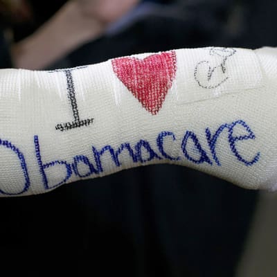 Henkilöllä on kädessään kipsi, johon on kirjoitettu englanniksi: "Rakastan Obamacarea". Rakastaa-verbi on korvattu punaisella sydänsymbolilla.