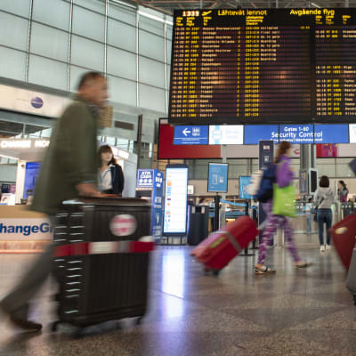 Resenärer med kappsäckar går på Helsingfors-Vanda flygplats. I mitten syns avgående flyg på en tavla.