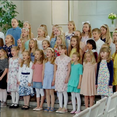 Pastellklädda barn med blommor i håret sjunger framme i en kyrka.