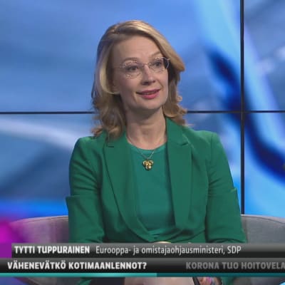 Europa- och ägarstyrningsminister Tytti Tuppurainen klädd i grönt ser in i kamera och ler. Hon sitter i Yles tv-studio där bakgrunden är blå och abstrakt.