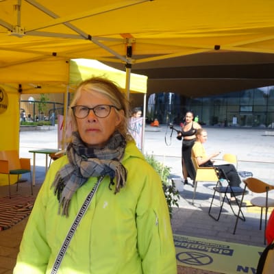 Eeva Kaila i en ljusgrön rock och halsduk inne i ett gult tält. Hon har glasögon på sig.  
