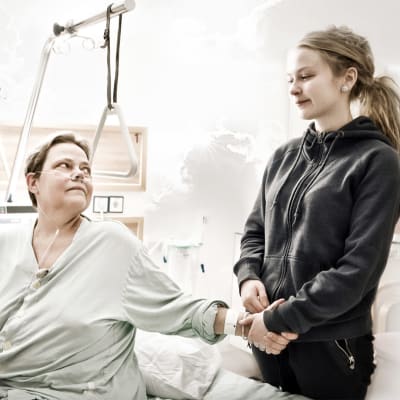 En mor sitter på en sjukhussäng och håller en vuxen dotter i handen.