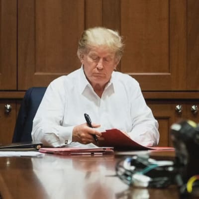 Donald Trump istuu pöydän ääressä sairaalassa ja katsoo papereitaan