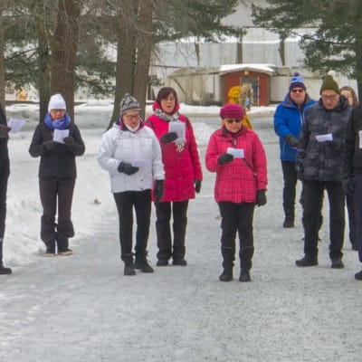 Ryhmä kuorolaulajia harjoittelee laulua lumisessa puistossa toppatakit yllään 