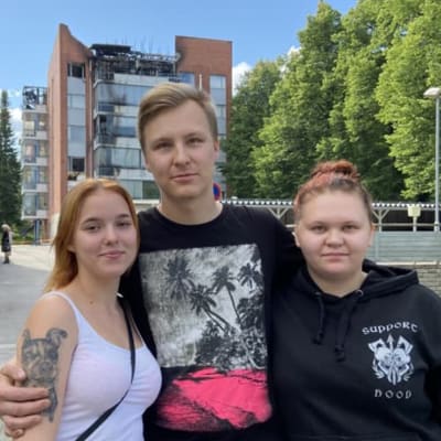 Eerika Pasanen, Samu Silventoinen och Sari Veijalainen står framför ett flervåningshus som brunnit 