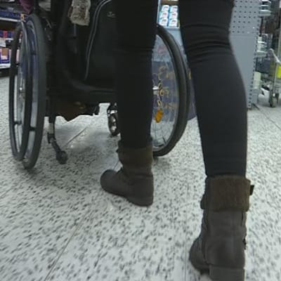 Henkilöhtainen avustaja työntää avustettavan pyörätuolia kaupassa.