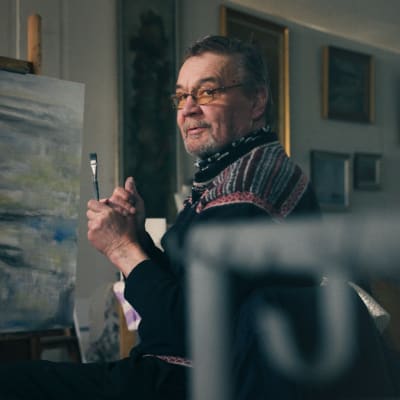 Veli Seppä katsoo kameraan hieman hymyillen pensseli kädessään edessään keskeneräinen maalaus.