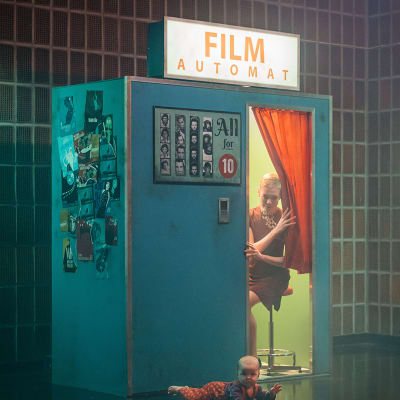 Film Automat -kyltti kopin katolla, nainen kurkistaa kuvauskopin verhon takaa, edessä vauva ryömii. Kuvassa teksti "Teeman elokuvafestivaali 2021"