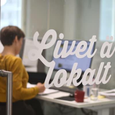 En kvinna jobbar vid sin dator bakom en glasdörr. På dörren står "Livet är lokalt".