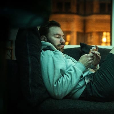 En man ligger på soffa och tittar på sin telefon. 