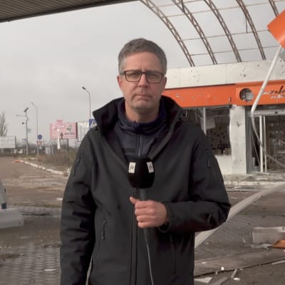 En man med en mikrofon i handen står på en förstörd bensinstation.