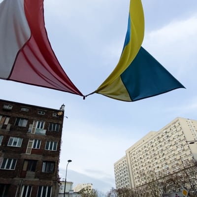 Polens och Ukrainas flagga vajar mot en blå himmel. I bakgrunden finns flervåningshus i Warszawa.