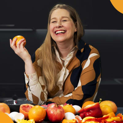 Kirjailija Leena Parkkinen nauraa appelsiini kädessään ja röykkiö vihanneksia ja hedelmiä edessään pöydällä.