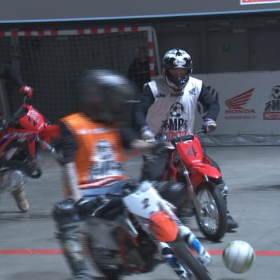 Neljä pelaajaa minicrosspyörien selässä tavoittelee jalkapalloa. Motoball-pelaajilla motocross-kypärät päässä. Ajoasuen päällä joukkueiden kevytliivit: valkoiset ja oranssit.