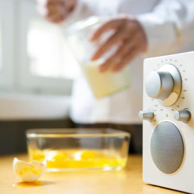 Keittiönpöydällä etualalla radiovastaanotin, jonka vieressä kananmunankuoria. Ihmishahmo mittaa taustalla sokeria, aikoo leipoa jotain.