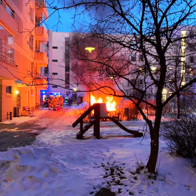 Personal från räddningsverket står i grupp bredvid en lekstuga som brinner mellan en mängd bostadshus.