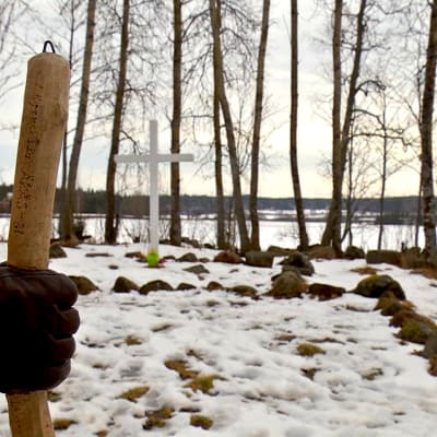 En hand som håller i en vandringskäpp. I bakgrunden ett träkors. 