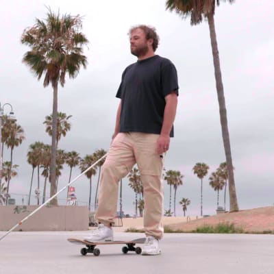 Brian Bushway seisoo betonisella aukiolla toinen jalka rullalaudan päällä kädessään valkoinen keppi. Taustalla on Venice Beach ja palmupuita.