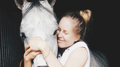 En ung kvinna kramar med slutna ögon sin hästs mule. Hästen är silvergrå.