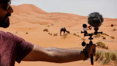 En man håller i en filmkamera mot en kamel i öknen