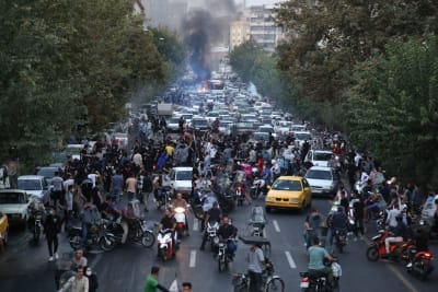 Demonstration i Teheran. Människor, bilar och mopeder täpper till en trädkantad gata. Vid ändan av gatan syns byggnader och nära dem brinner något på gatan.