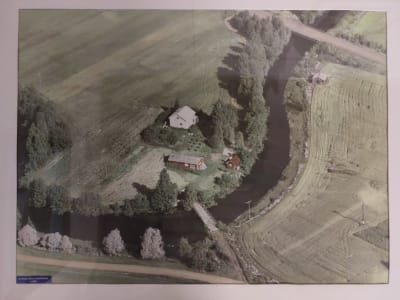 Ilmakuva peltojen ja vuolaan joen ympäröimään niemeen, jossa on valkoiseksi maalattu talo ja piharakennuksia.