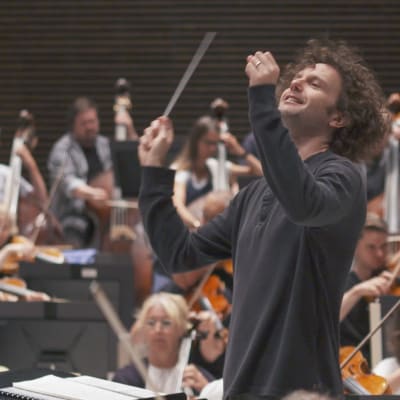 Radion sinfoniaorkesterin hymyilevä ylikapellimestari Nicholas Collon johtaa RSO:n harjoituksia Musiikkitalon Paavo-harjoitussalissa pukeutuneena mustaan puuvillapaitaan, Orkesterin soittajia epätarkkoina taustalla.