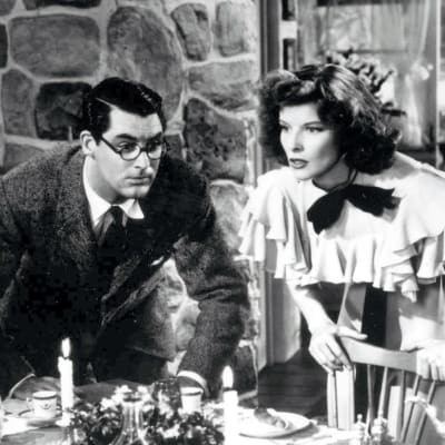 Cary Grant ja Katharine Hepburn elokuvassa Hätä ei lue lakia: molemmat seisovat päivällispöydän ylle kumartuneina, Grant tuijottaa hieman hölmistyneen näköisenä vastapäätä olevaa naista ja Hepburn on sanomassa jotain vastapäätä itseään olevalle miehelle.