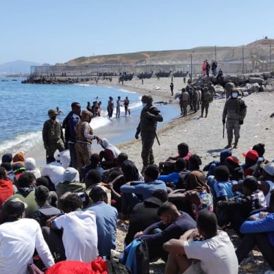 Suuri joukko siirtolaisia istuu rantahiekalla. Rannalla seisoo sotilaita varusteissaan, kasvomaskit päällä.