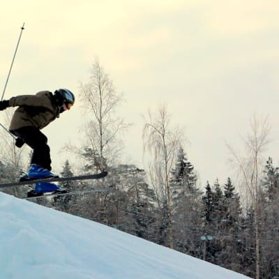 En pojke med brun jacka och svart hjälm hoppar med sina skidor över ett gupp i en slalombacke.