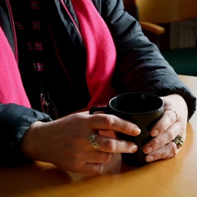 En kvinna sitter vid ett bord med en kaffekopp i händerna. Kvinnans ansikte syns inte på bilden.