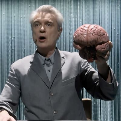 Harmaapukuinen David Byrne laulaa puolikuvassa lavalla aivot kädessä