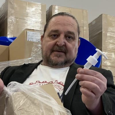 En man håller i en liten plastpump och en låda fyllda med sådana i en lagerhall med pafflådor. 