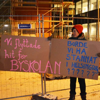 En kvinna står utomhus och håller i två skyltar där det står: Vi flyttade hit för byskolan borde vi ha stannat i Helsingfors?