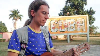 Man spelar gitarr utomhus framför stor skylt med texten "Cuba"
