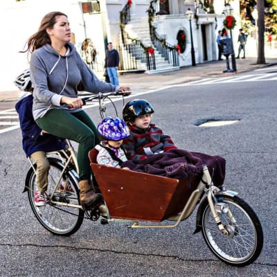 En kvinna cyklar men två barn sittandes fram i en korg och ett barn på pakethållaren.