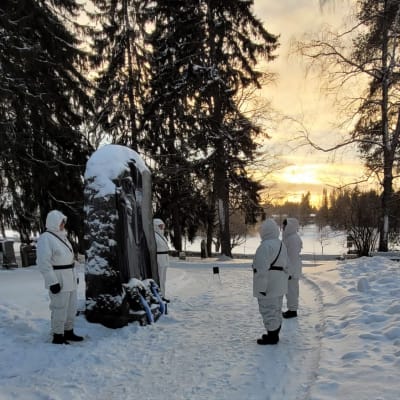 Lumipukuiset miehet seisovat kivisen muistomerkin molemmilla puolilla talvipäivänä.