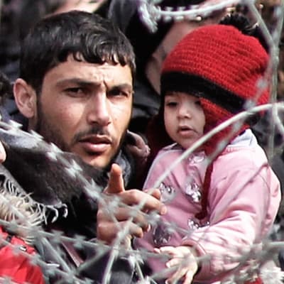 Joukko Kreikkaan pyrkiviä turvapaikanhakijoita seisoo piikkilanka-aidan takana. Kuvan keskellä on parrakas mies, joka pitelee sylissään pikkulasta ja osoittaa sormellaan kuvaajan suuntaan.*
