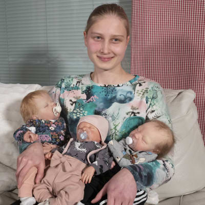 Elviira Närvä istuu sohvalla sylissään kolme aidon vauvan näköistä Reborn-nukkea.