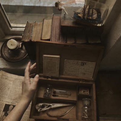 Gamla böcker, tidningar och en låda fylld med små saker. En hand som rör vid lådan och ett ljus syns i bakgrunden.