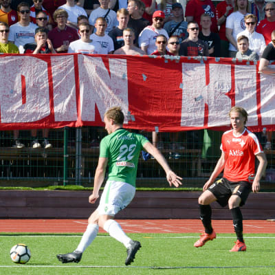 En bild på en fotbollsspelare i grönvitt (EIF) som sparkar på en boll. Bredvid honom finns spelare i rött och svart. Bakom fotbollsspelarna finns en banderoll där det står stadin kingit - de stöder laget i rött och vitt (HIFK)
